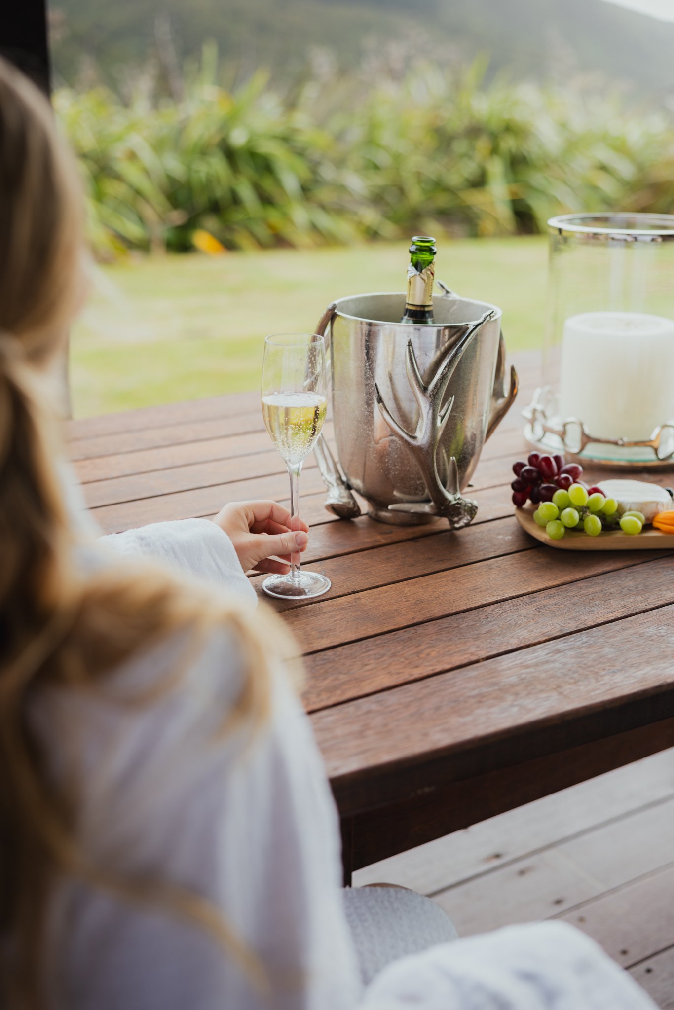 A person holding a champagne flute with a glass of champagne on a wooden table, with a bottle in an ice bucket, a plate of grapes, and a candle, outdoors with greenery in the background.