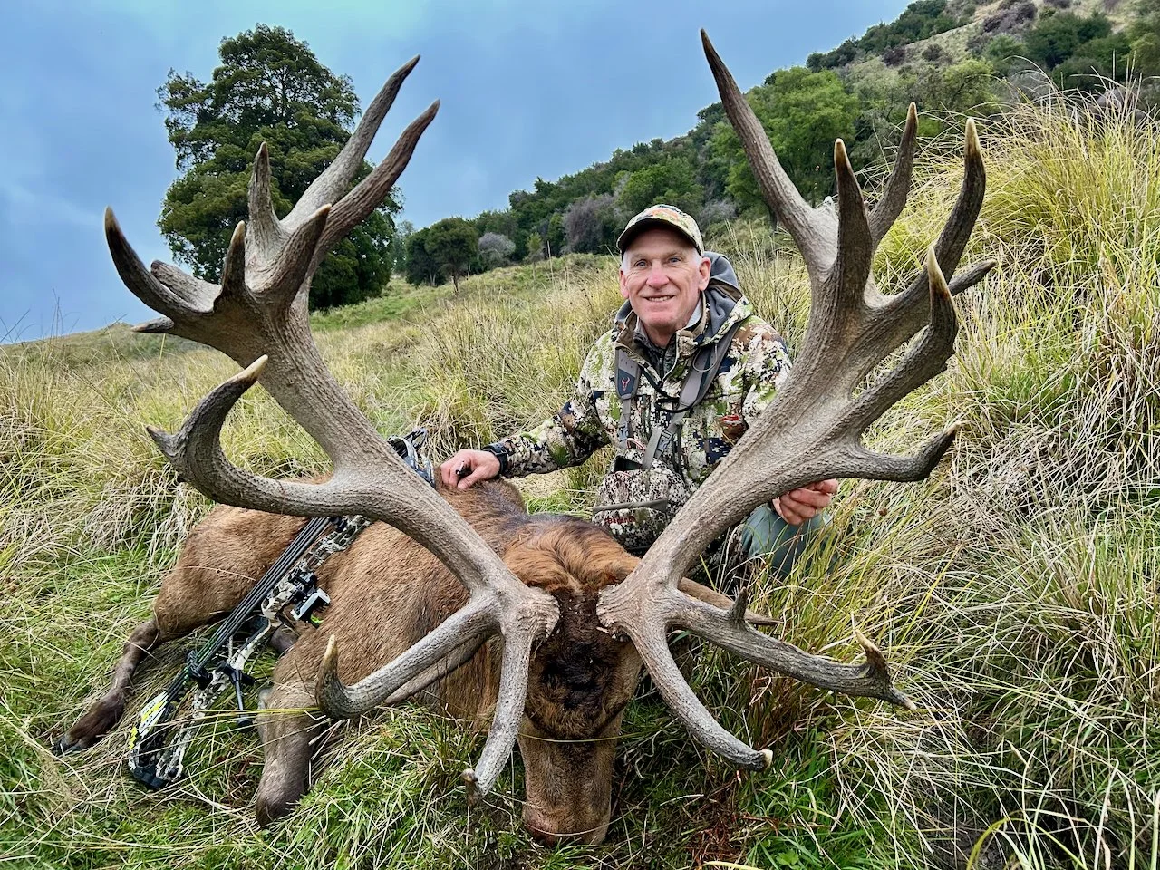 A man in camouflage clothing kneels behind a large elk he has hunted, holding its antlers in a grassy field with trees and hills in the background.