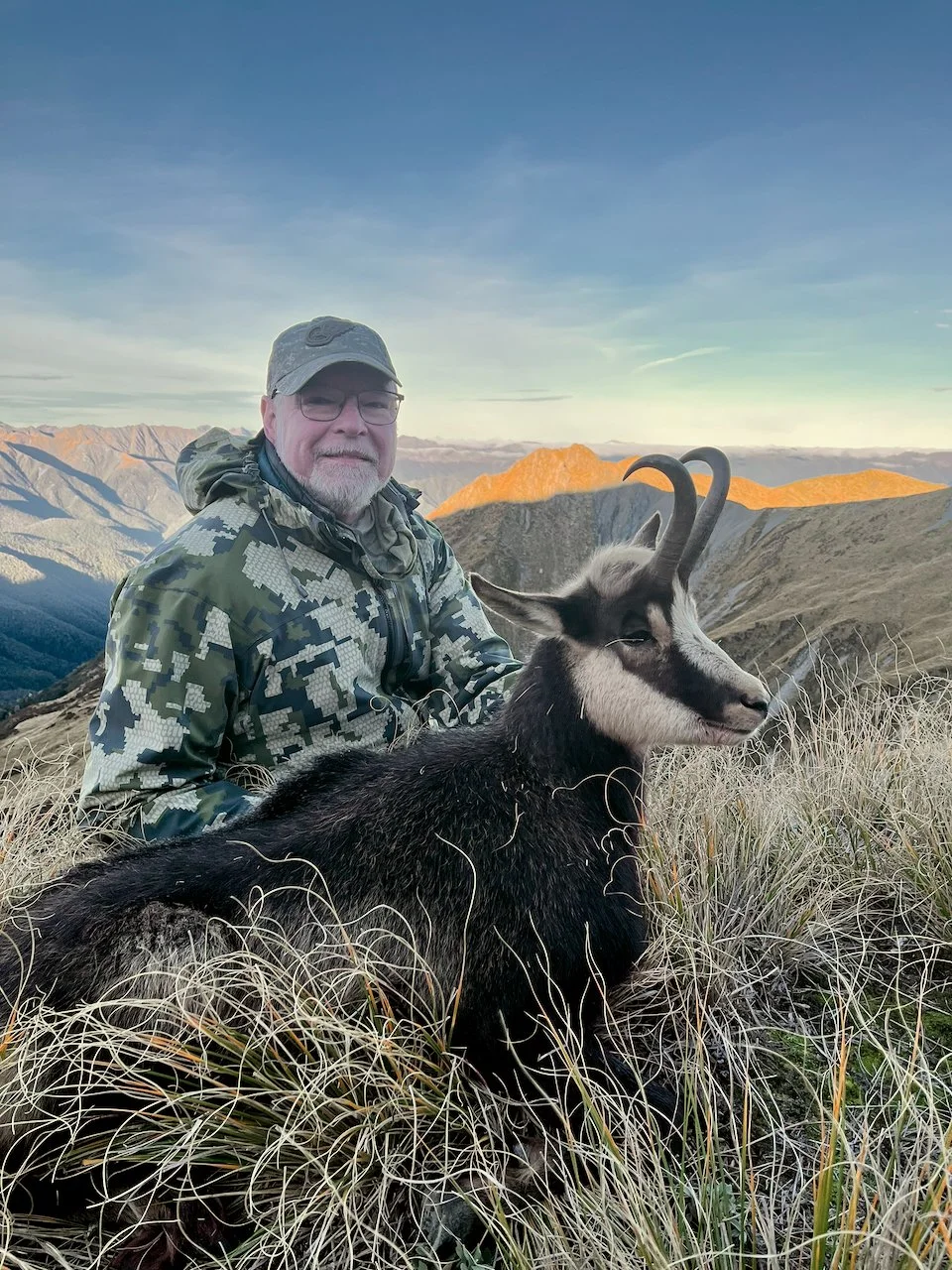 A man in camouflage jacket and cap sitting on grass with a black and white goat with curved horns against a mountain landscape at sunrise or sunset.