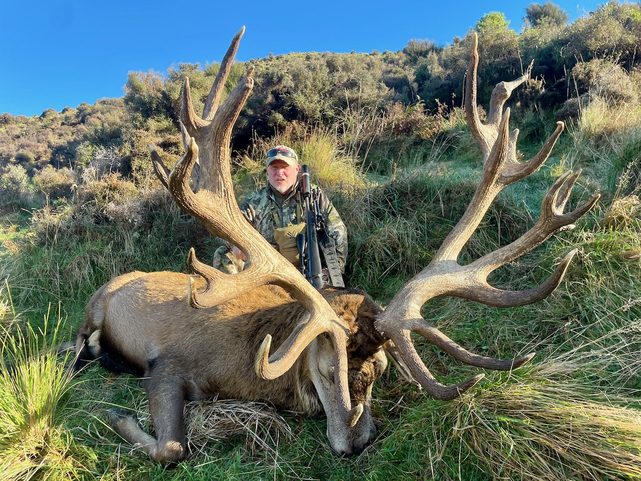 Hunter in camouflage clothing kneeling behind a large elk with impressive antlers, outdoors in a grassy, hilly area under a clear blue sky.