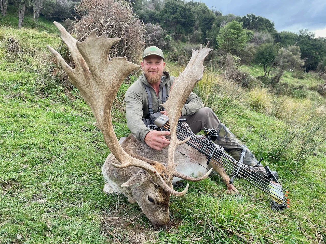 Man sitting on grass with a bow, posing next to a large deer he has hunted, in a green outdoor landscape with trees and shrubs.