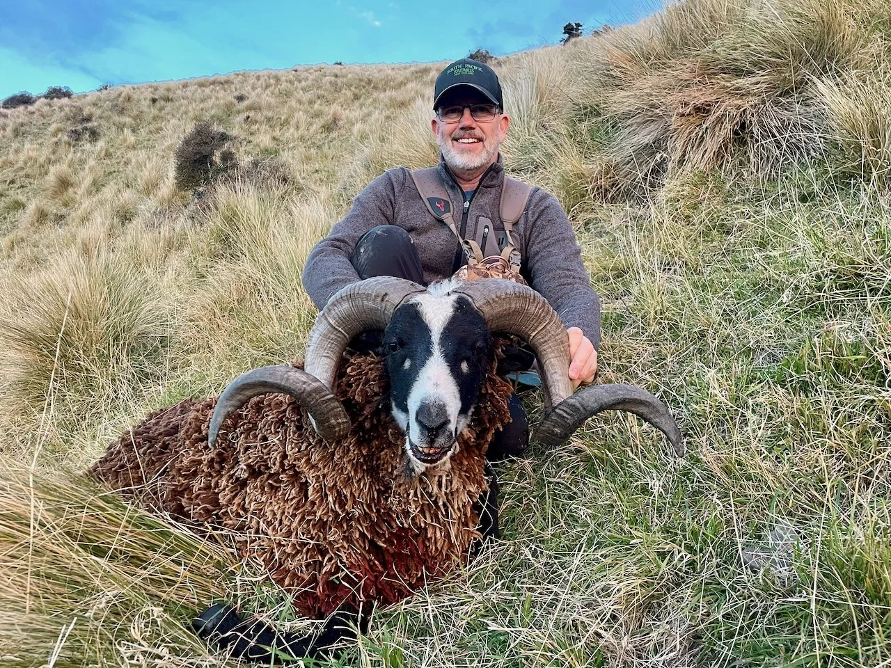 A man with glasses, a beard, and wearing outdoor gear is sitting on a hillside grassy area, holding a large sheep with prominent curved horns. The sheep has a thick, curly brown coat and black and white facial markings.