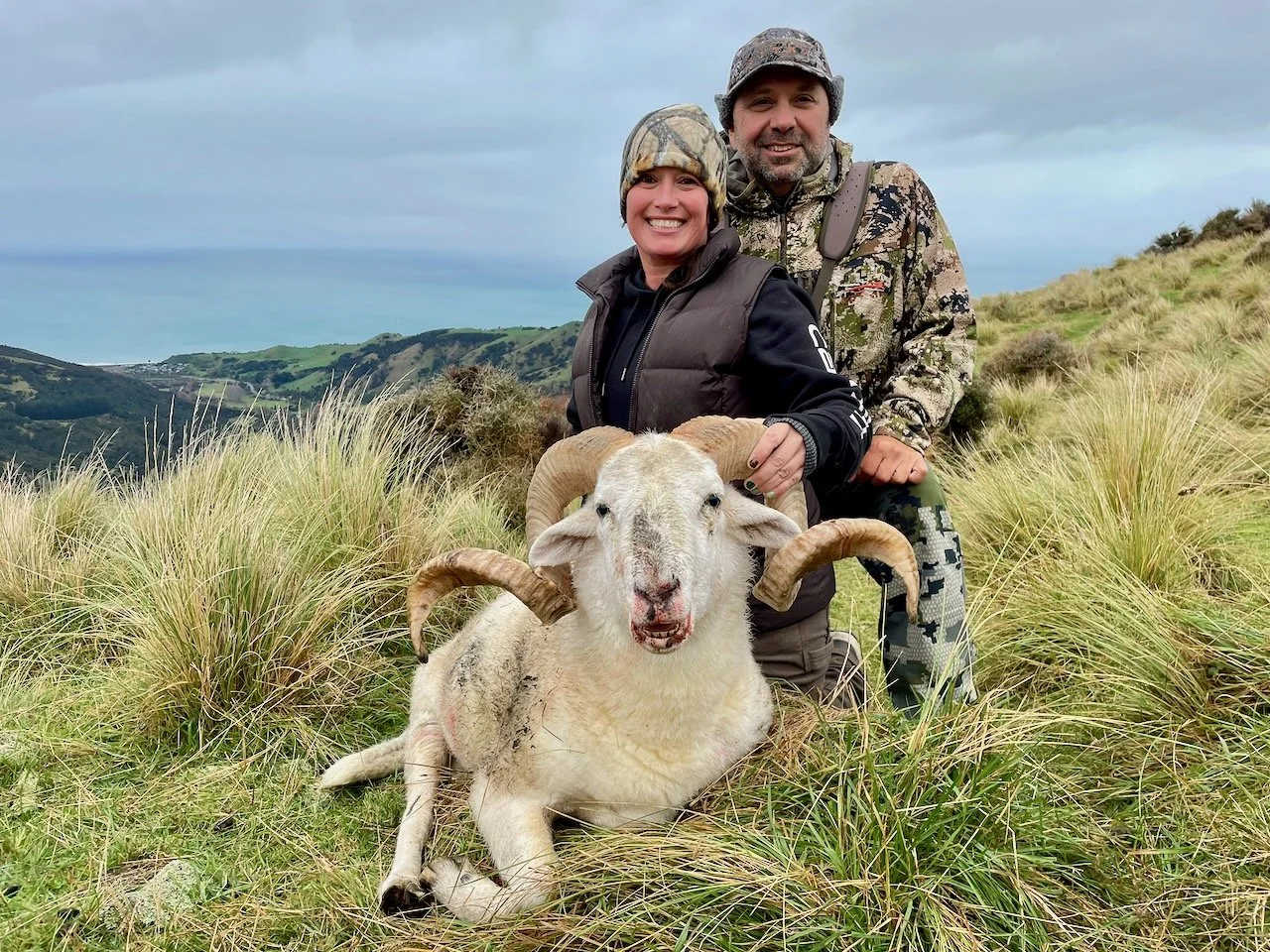 A smiling woman and man in outdoor gear kneel behind a sheep with large curved horns in a grassy landscape with rolling hills and cloudy sky.