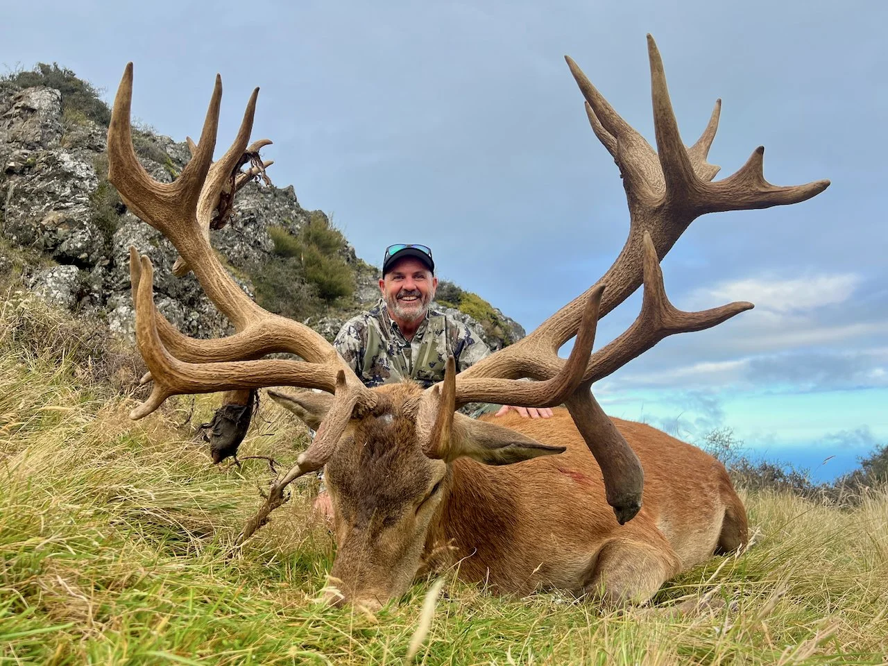 A man with a big smile wearing outdoor gear and sunglasses kneels behind a large elk with impressive antlers, lying in tall grass in a mountainous area under a partly cloudy sky.
