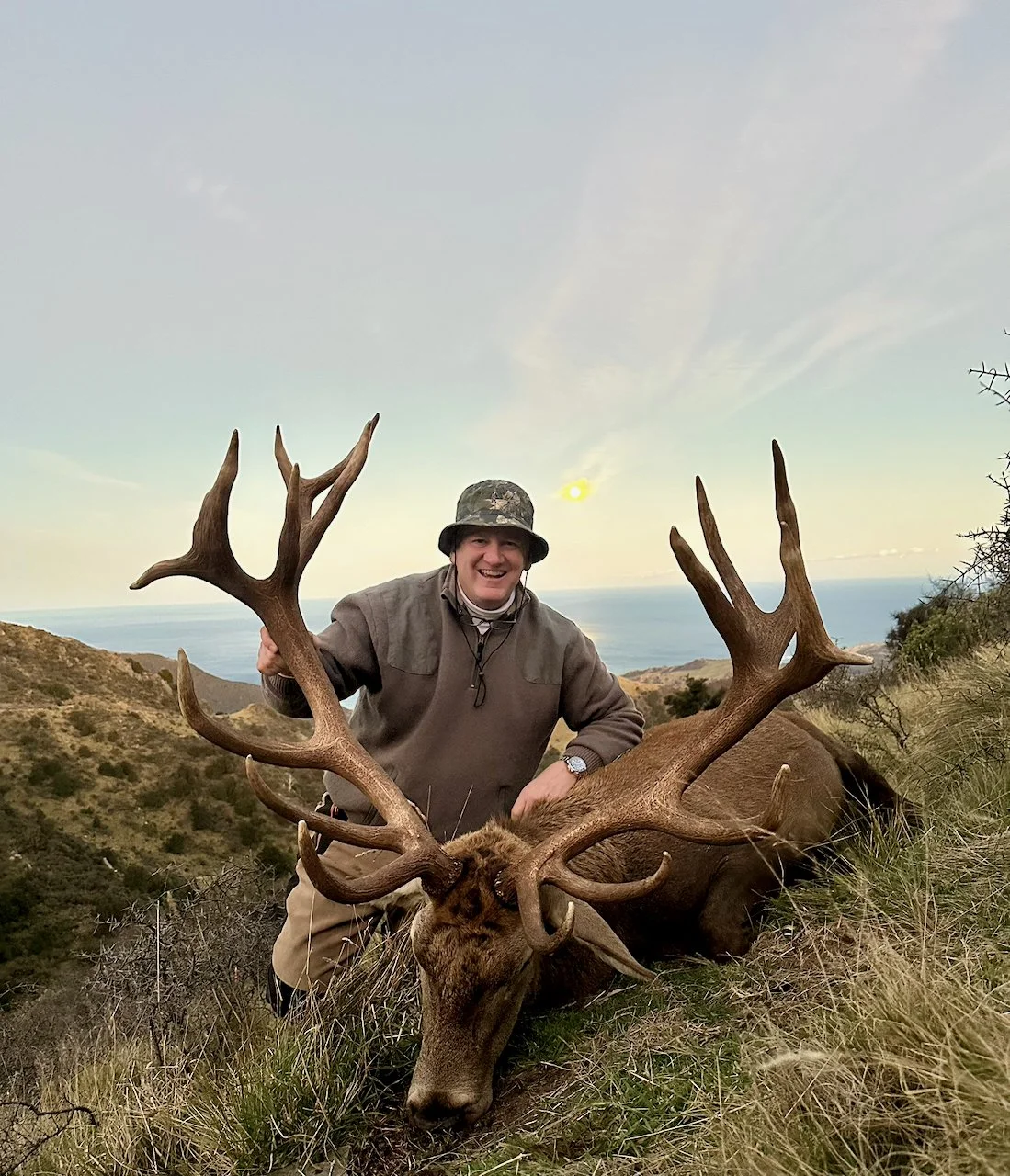 A smiling man wearing outdoor clothing and a camouflage hat posing behind a large elk with big antlers in a hilly landscape near the coast.