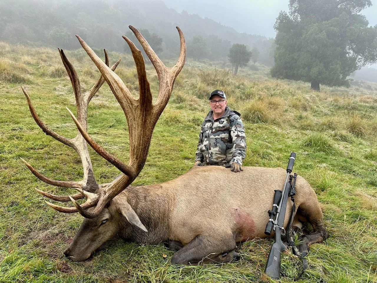 Man in camouflage clothing kneeling next to a large dead elk with impressive antlers. A rifle is resting on the elk's side on a grassy field with misty hills and trees in the background.