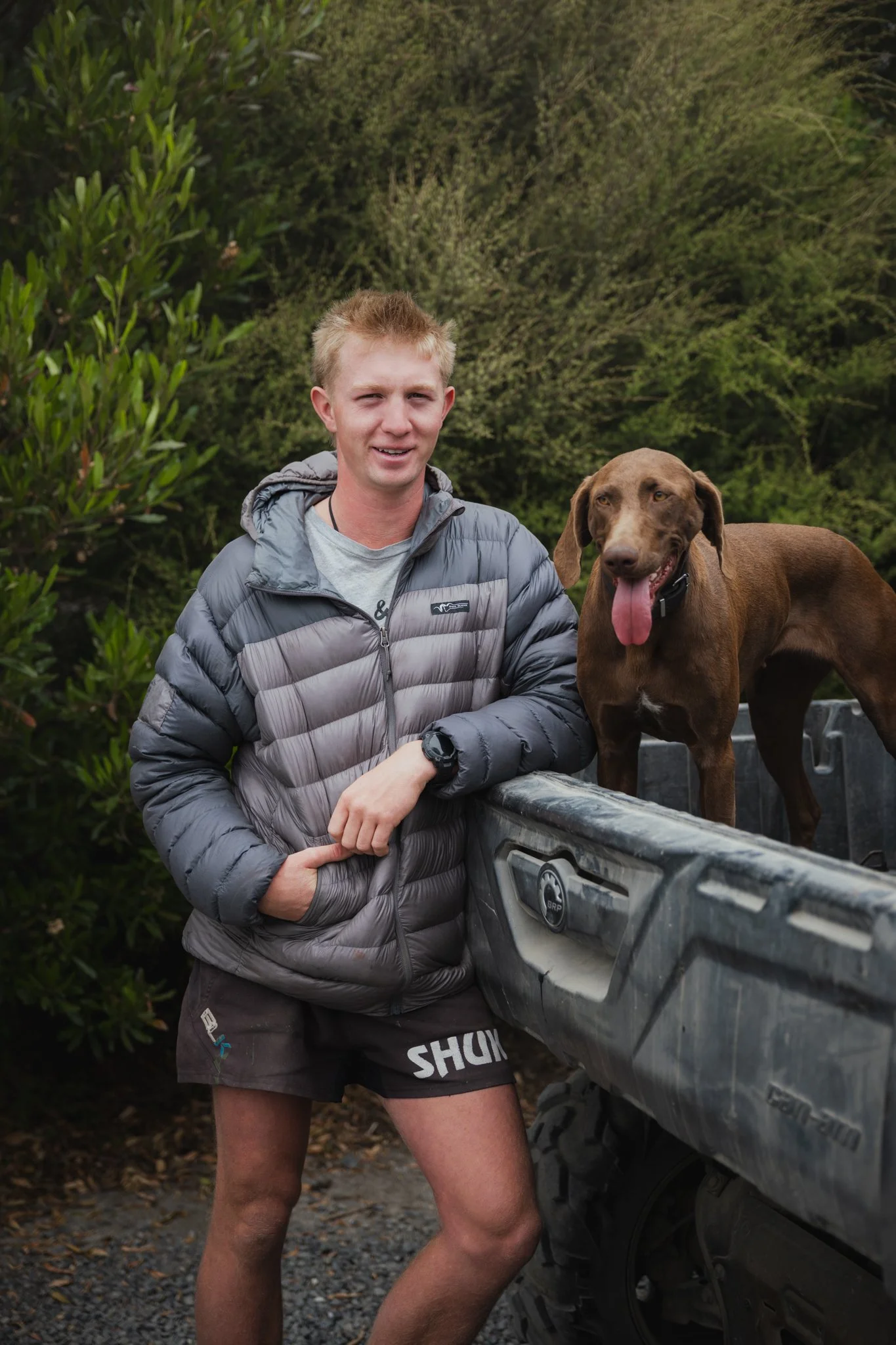 A young man is standing outdoors next to a truck, with a brown dog standing in the truck bed. The man is wearing a gray jacket and shorts, and the dog has its tongue hanging out.