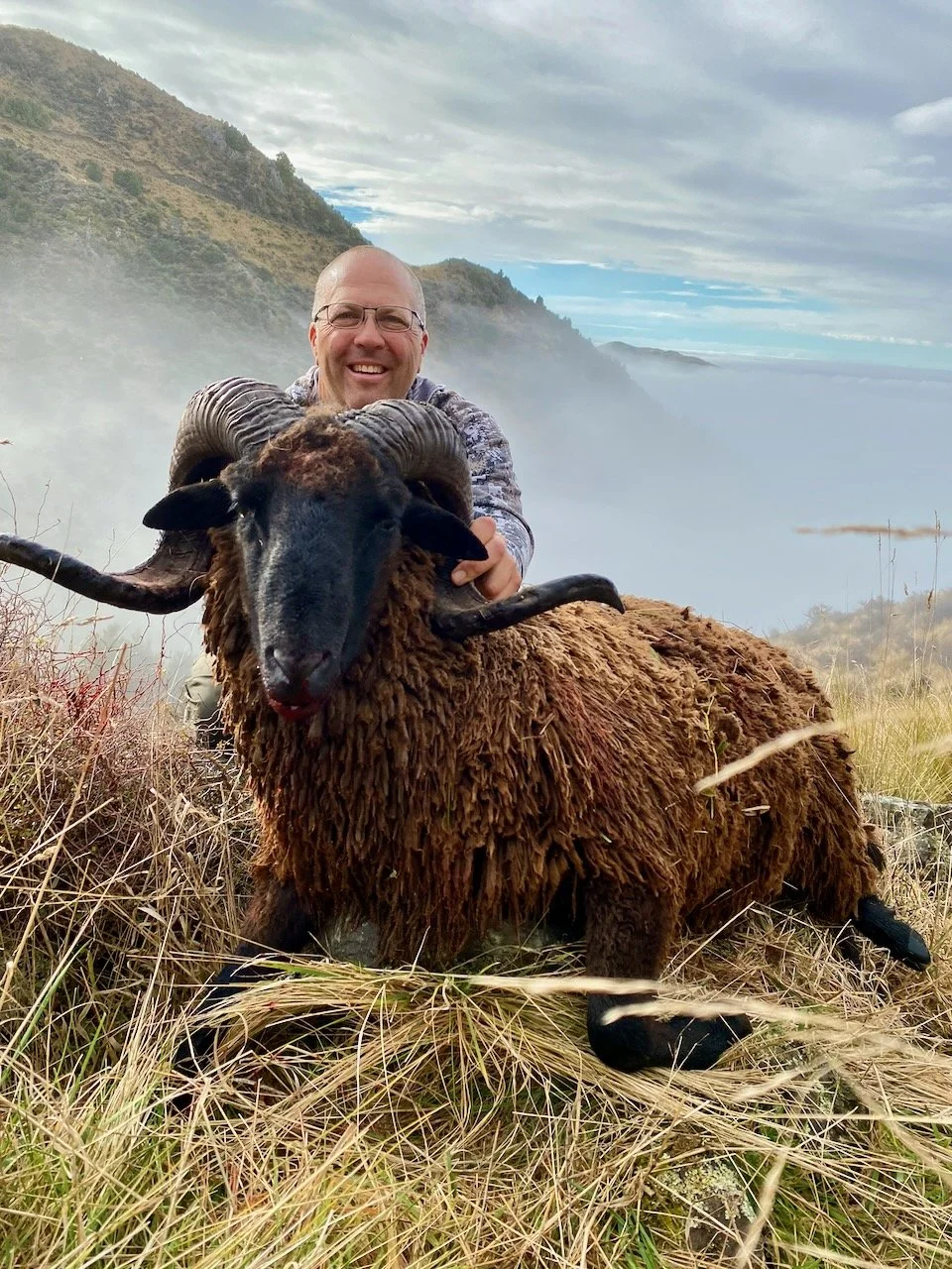 A man with glasses and a smile kneeling behind a large, brown, woolly sheep with curved horns, in a grassy field near a body of water with fog and hills in the background.