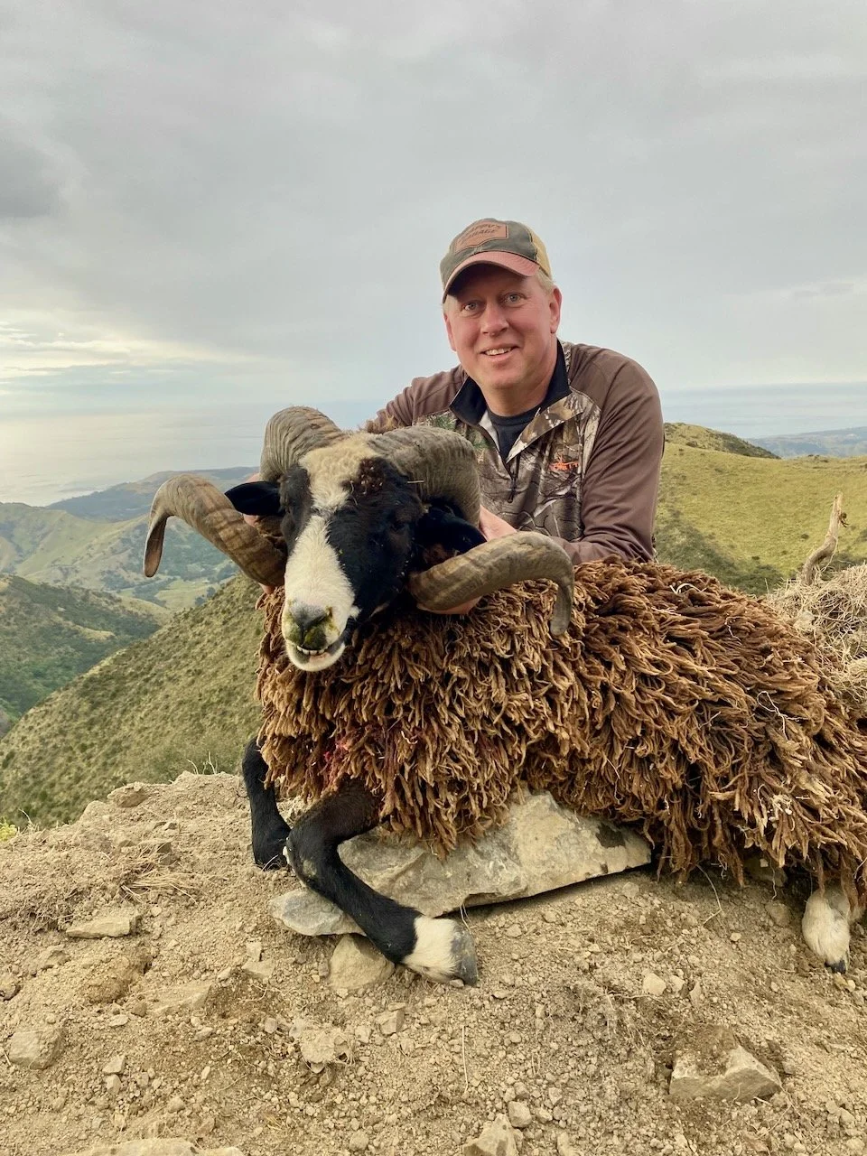 A man smiling outdoors on a mountain trail with a sheep that has large curved horns, a black face, and a brown woolly coat.