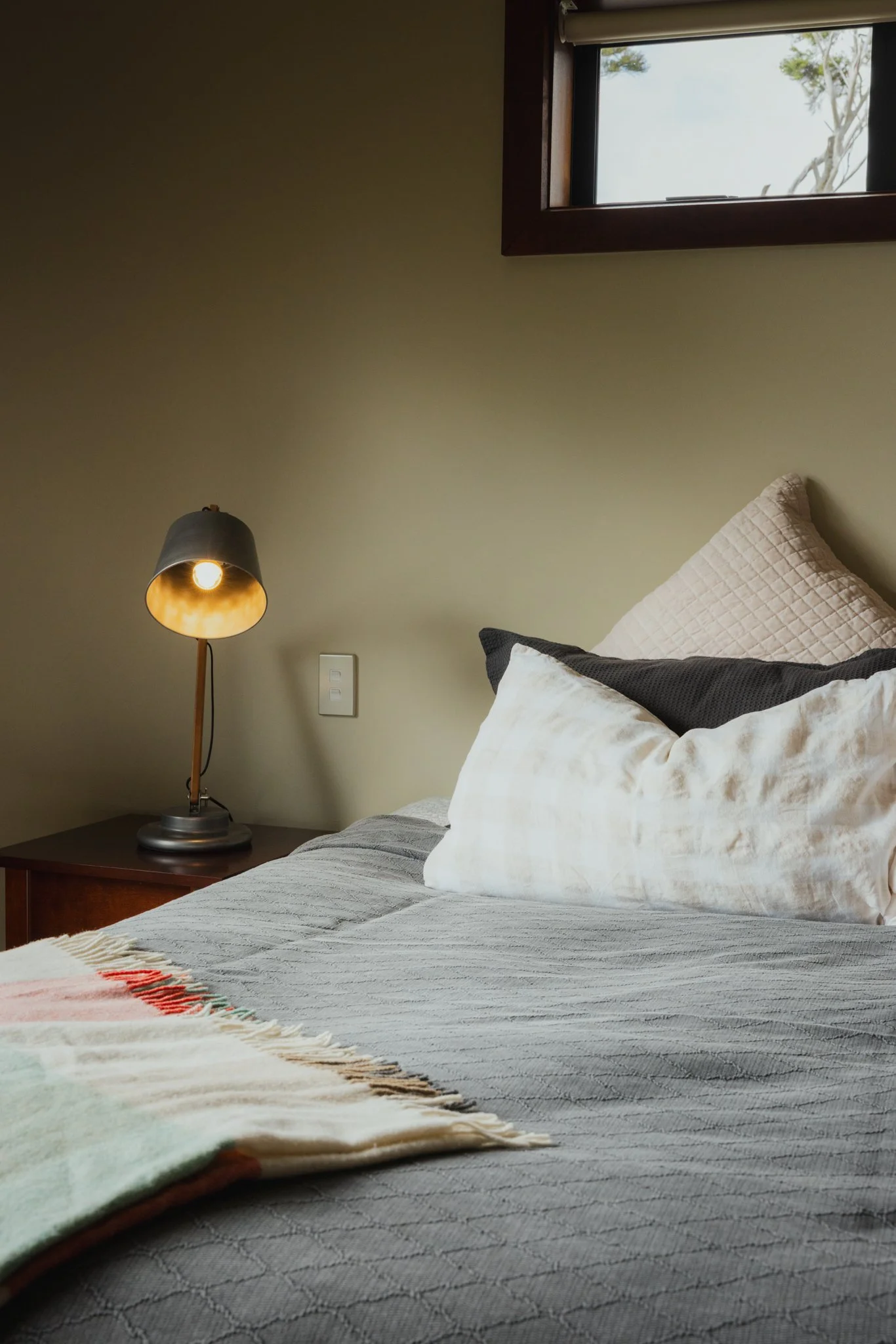 A bedroom scene with a neatly made bed, white pillows, a bedside table, a lamp turned on, and a window showing trees outside.