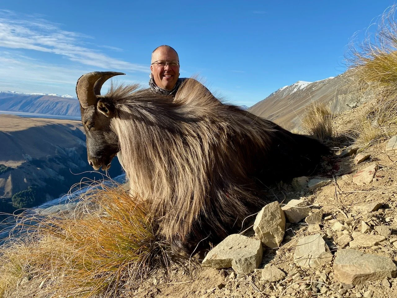 A man with glasses and a scarf smiling while sitting behind a large goat with curved horns on a rocky hillside with mountains in the background.