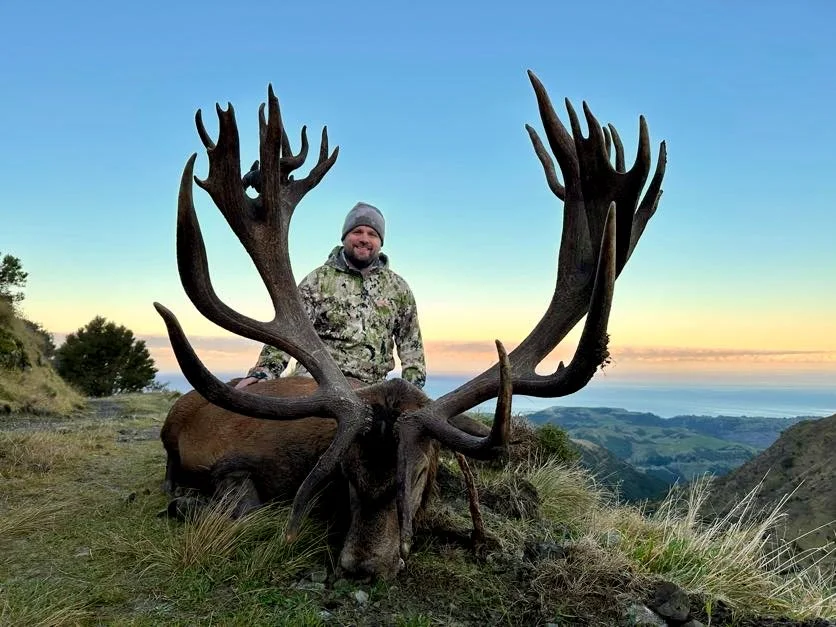 A man in camouflage clothing and a gray beanie sits behind a large elk with prominent antlers, on a grassy hillside with a scenic landscape of hills and water in the background during sunset.