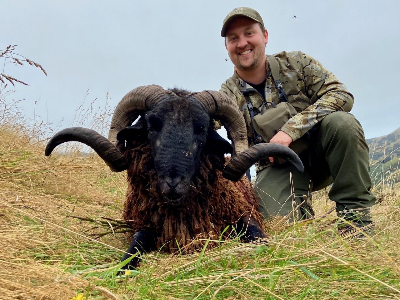 A smiling man in outdoor camouflage clothing crouches next to a large ram with curved horns in a grassy field.