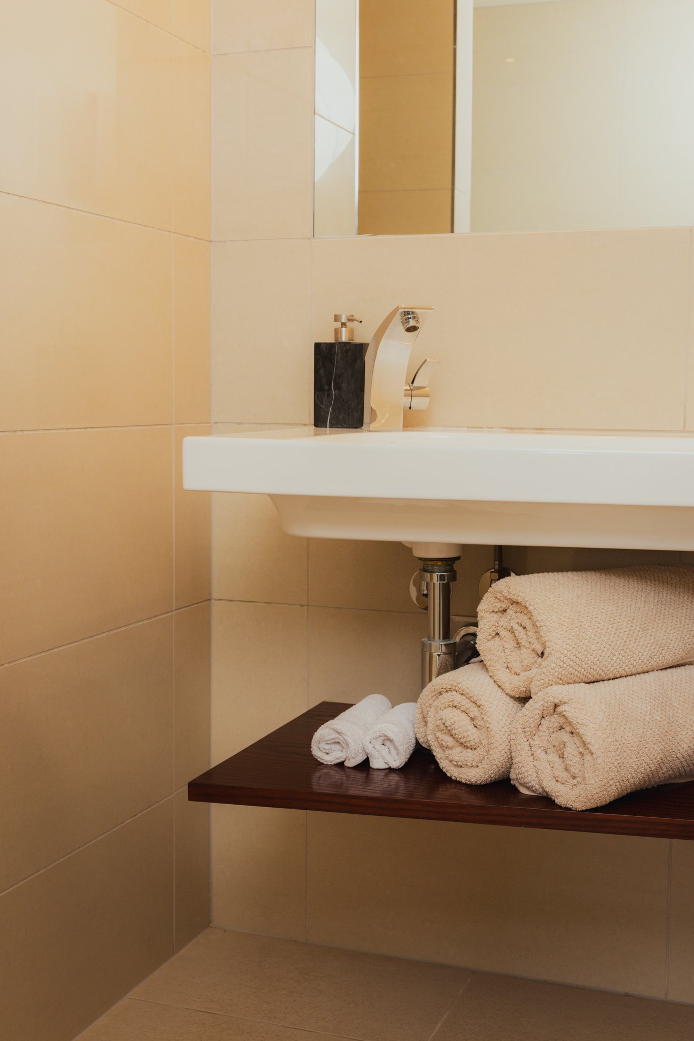 A bathroom sink with a faucet, a soap dispenser, a mirror, and a few rolled towels on a wooden shelf.