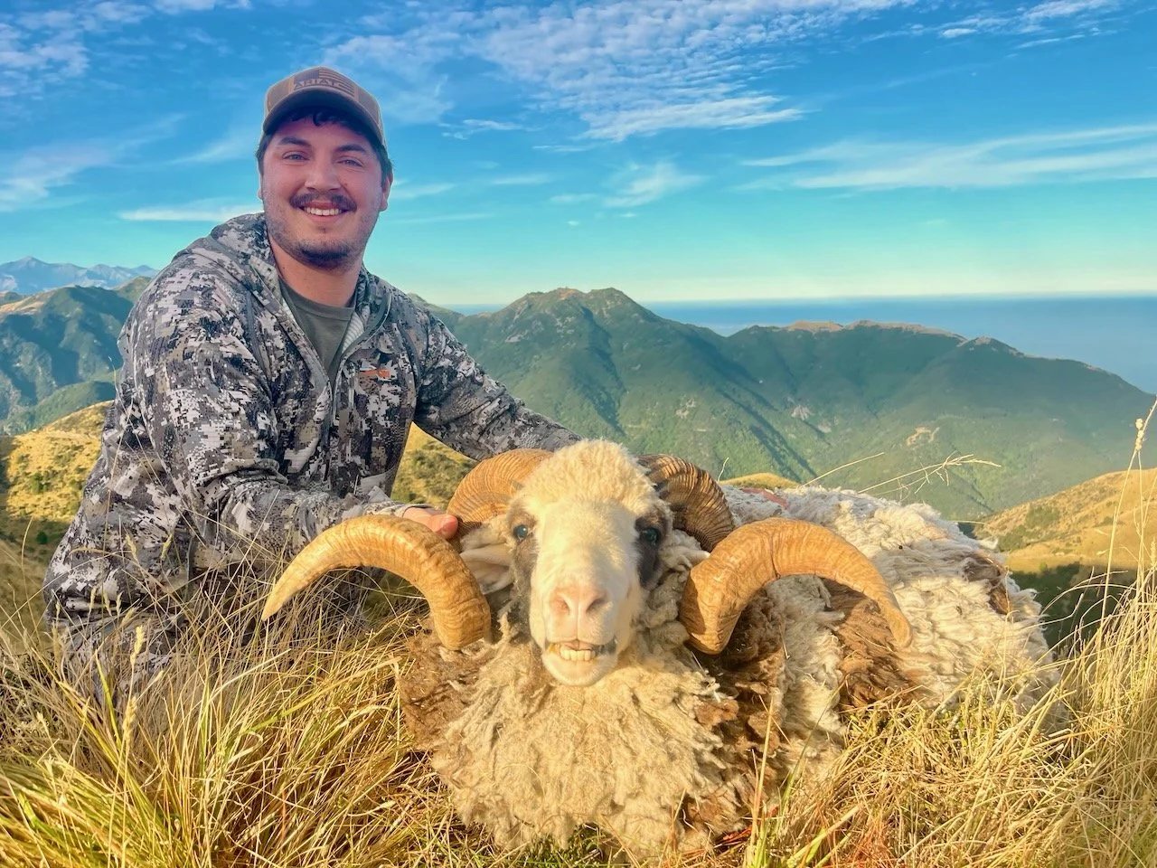 A man in camouflage clothing and a cap next to a sheep with large curved horns on a mountain trail, with a scenic mountain range and blue sky in the background.