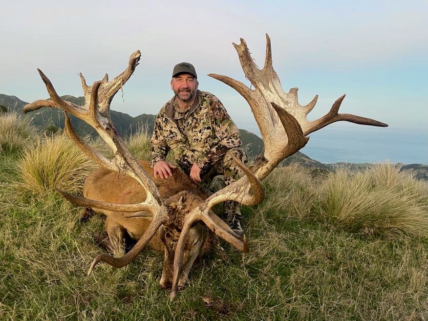 A man in camouflage clothing kneels behind a large, dead elk with impressive antlers in a grassy, mountainous landscape.