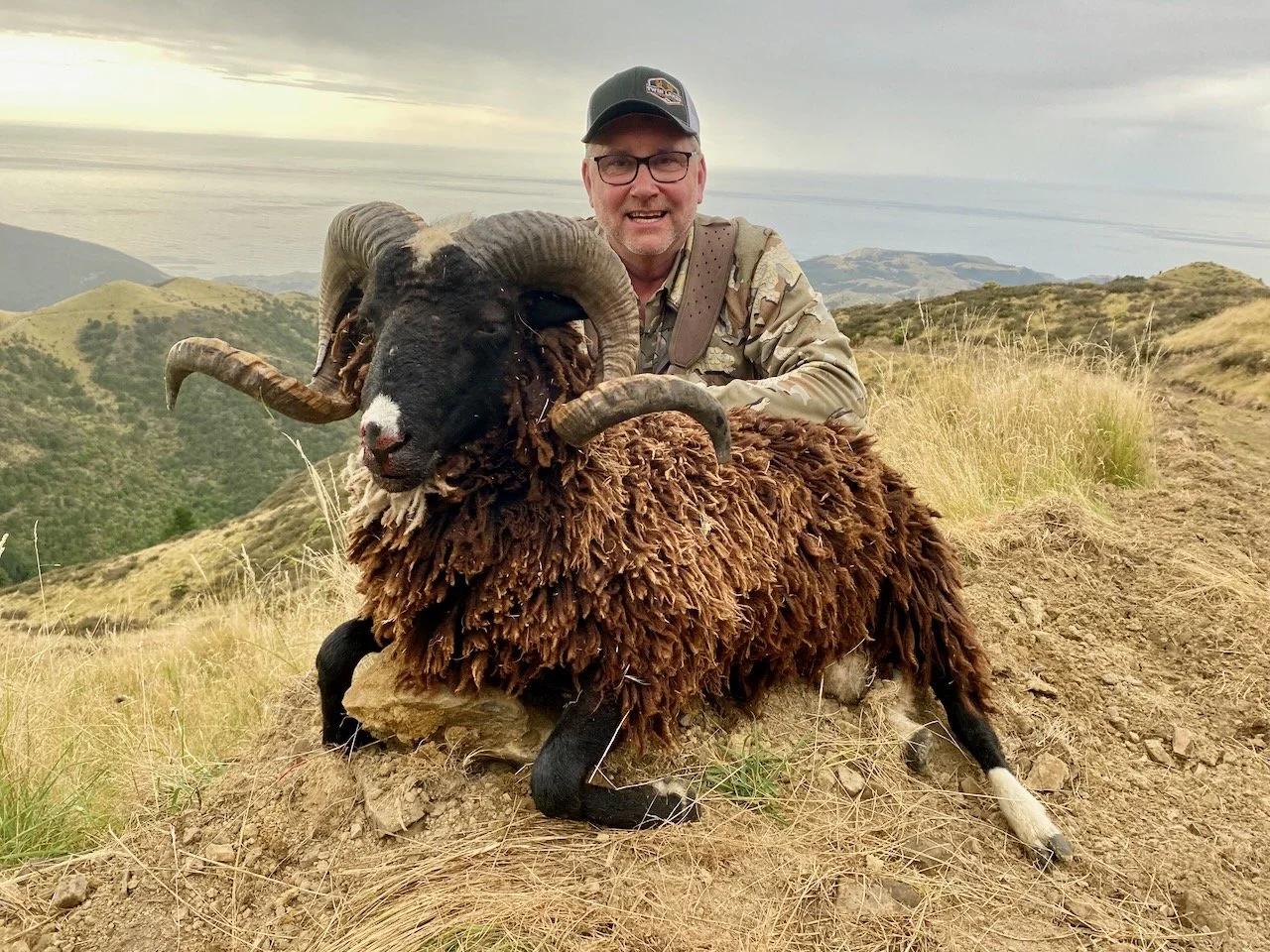 A man smiling and wearing a camouflage jacket and cap, posing with a large ram sheep with curled horns, on a grassy hillside overlooking a coastal landscape.