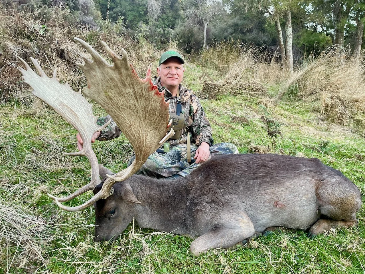 A man dressed in camouflage gear sits on grass next to a large dead elk with impressive antlers in a natural outdoor setting with trees and dry grass.