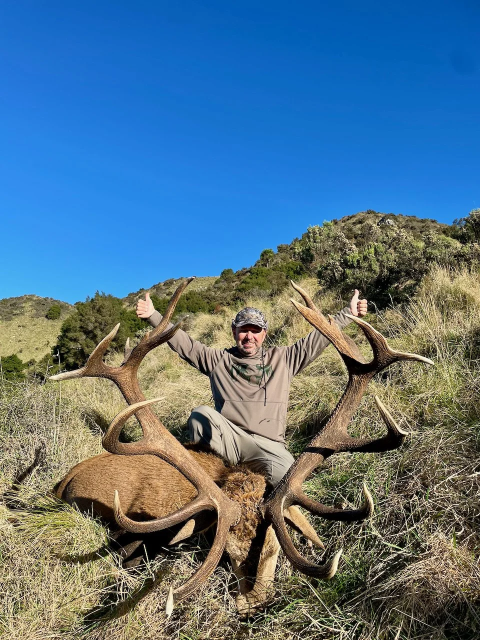 A man sitting on a large elk with impressive antlers in a grassy, hilly landscape underneath a clear blue sky, giving a thumbs up.