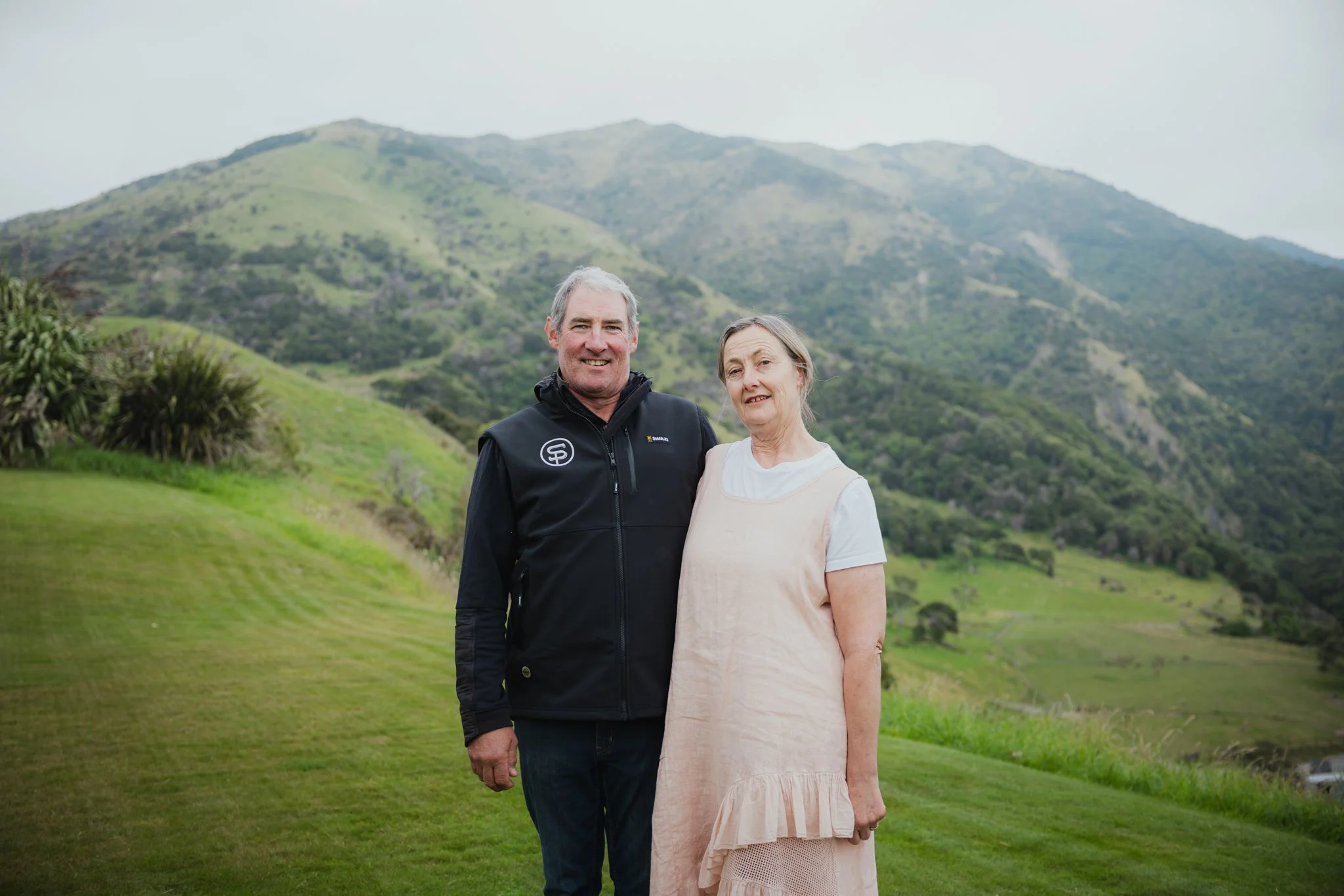 A middle-aged man and woman standing outdoors in front of a lush, green mountain landscape. The man is wearing a black jacket with a logo, and the woman is dressed in a light-colored dress. Both are smiling and looking at the camera.