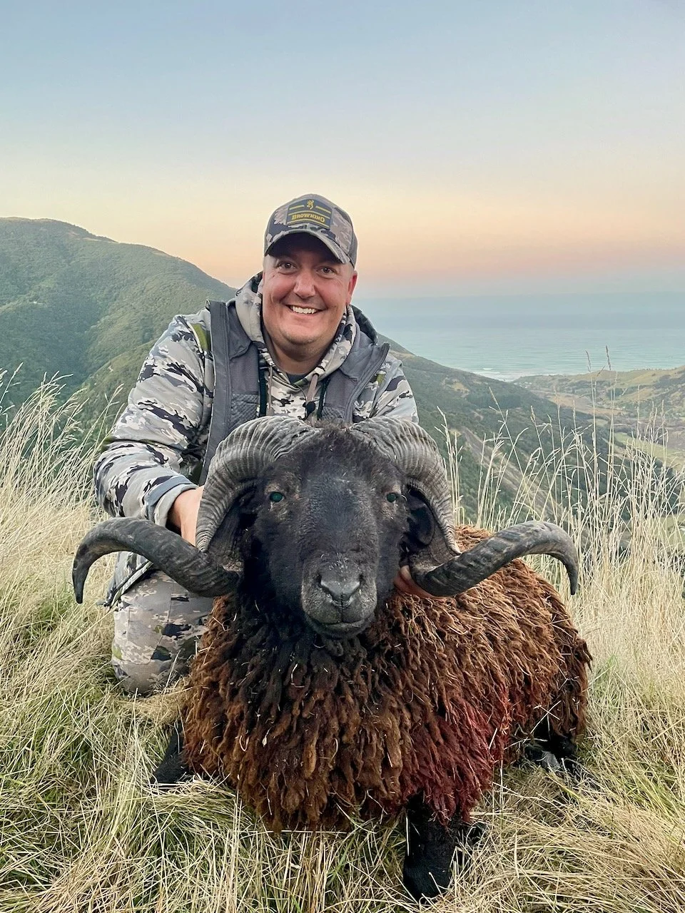 A man in camouflage clothing and a cap crouching in tall grass, smiling, while holding a large, black sheep with curved horns, in a hilly landscape during sunset.
