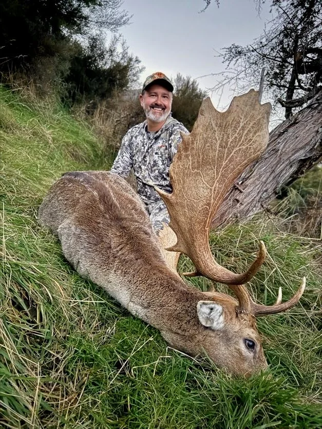 Man in camouflage clothing smiling next to a large fallen deer with antlers in tall grass and trees.