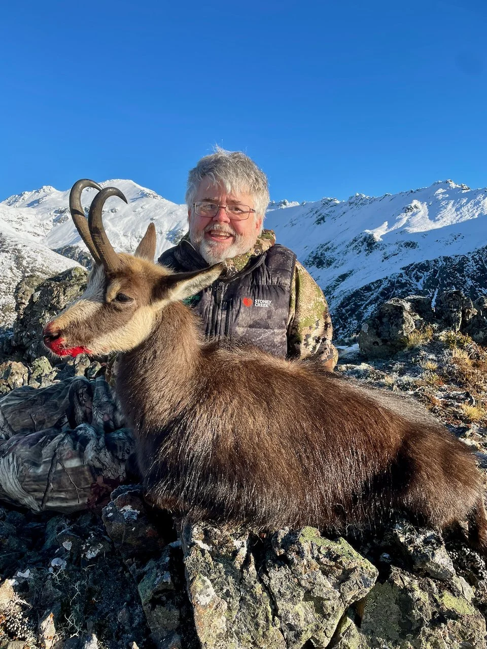 A man with gray hair and glasses smiling while sitting outdoors in a snowy mountainous landscape, with a tan and brown mountain goat with curved horns beside him.