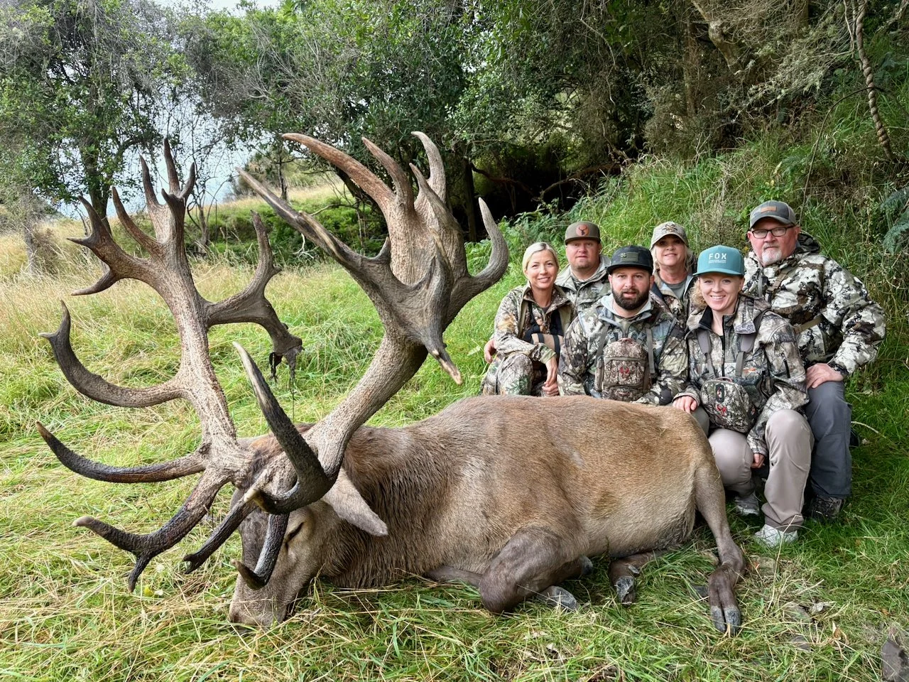 A group of six people posing outdoors next to a large, dead elk with impressive antlers, lying on the grass.
