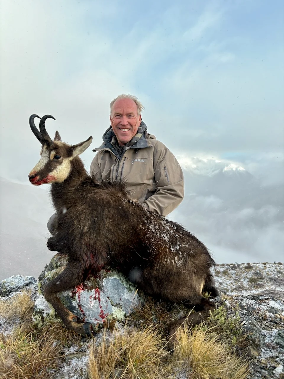 A smiling man in outdoor clothing kneels beside a dead mountain goat on a rocky and grassy terrain, with cloudy sky in the background.