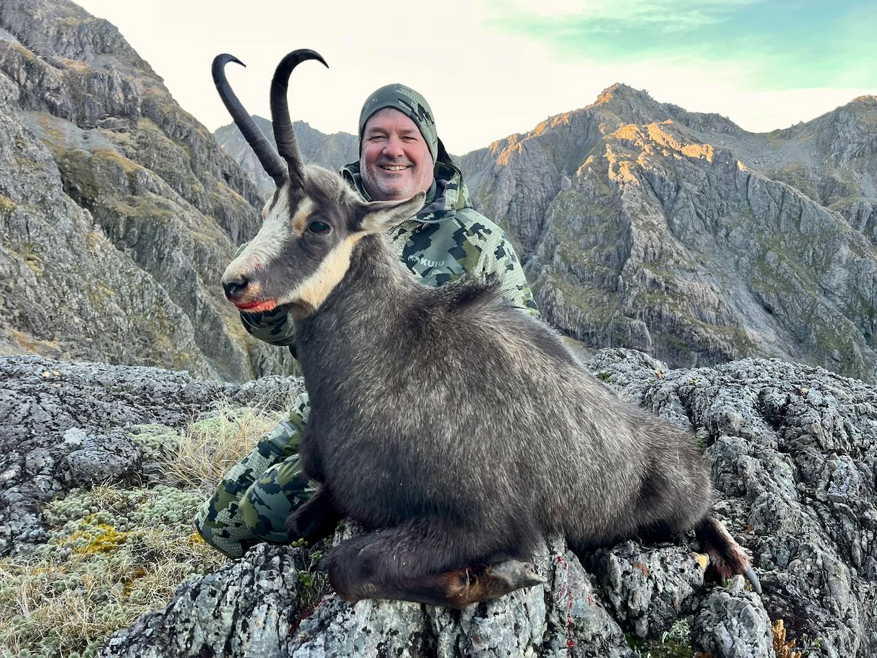 A smiling man in camouflage attire kneels behind a large goat with curved horns on rocky terrain in a mountainous landscape.