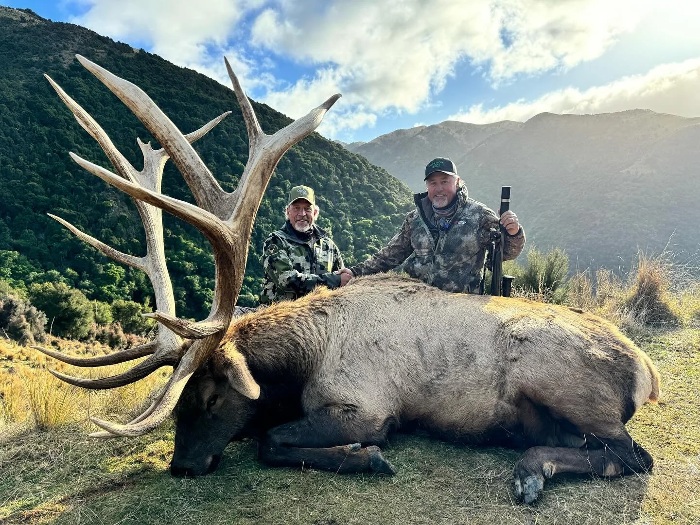 Two hunters in camouflage jackets shake hands next to a large, deceased elk with impressive antlers in a mountainous landscape.