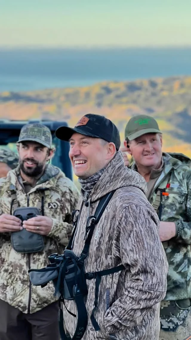 Three men in outdoor camouflage clothing standing outdoors with a scenic background of hills and water, smiling and looking happy.