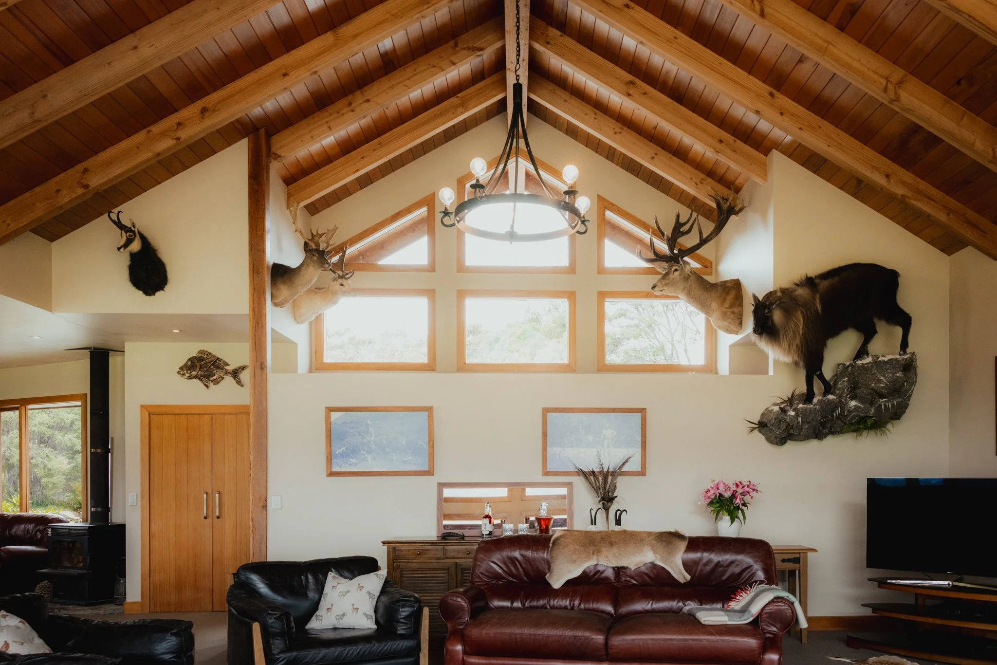Living room with wooden vaulted ceiling, hanging chandelier, and mounted animal heads with a window showing greenery outside.