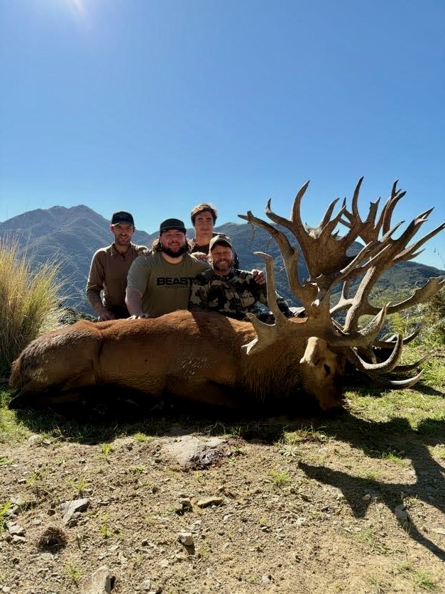 Four men standing behind a large dead elk with an expansive set of antlers, outdoors in a mountainous area under a blue sky.