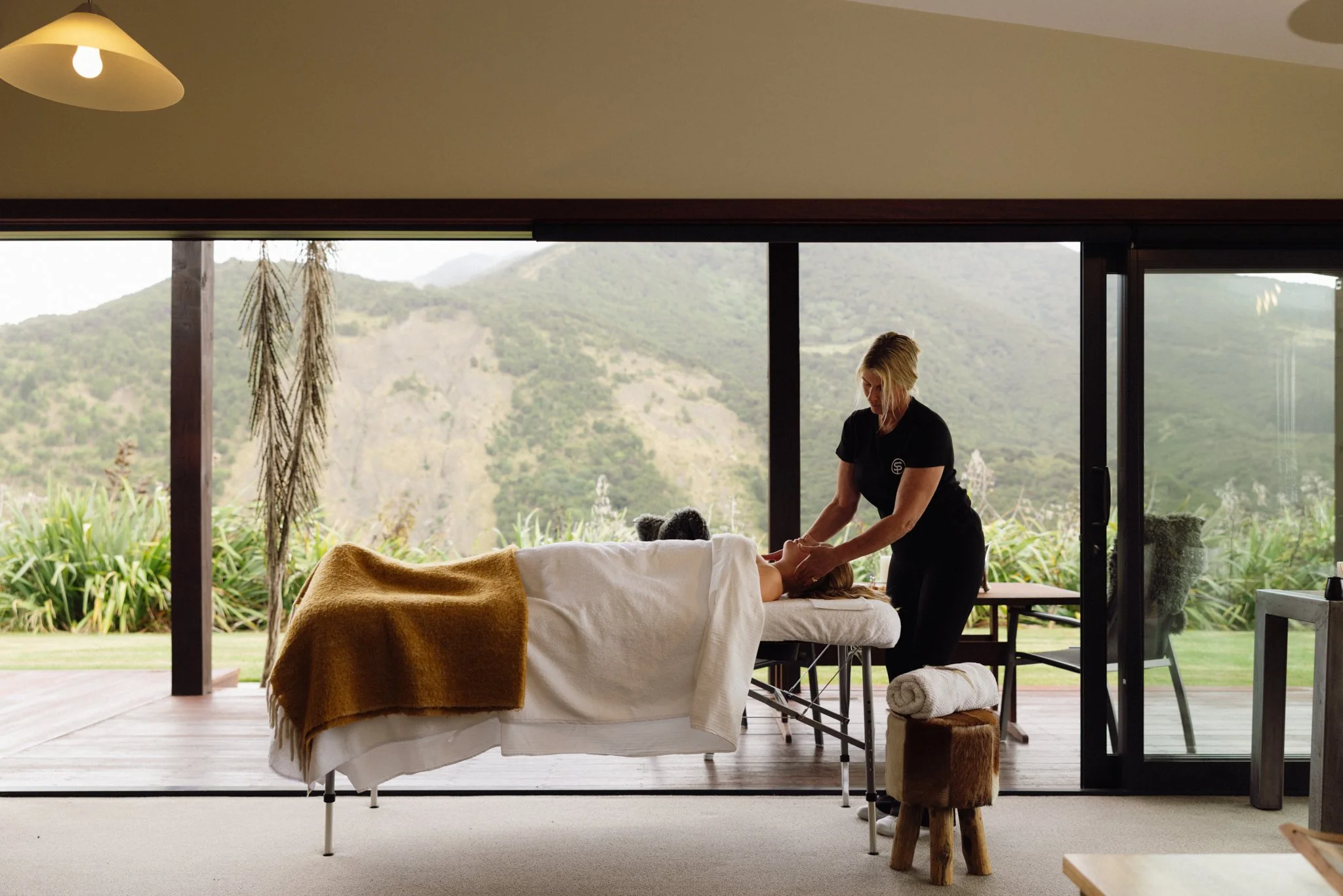 A person receiving a massage on a massage table inside a room with large open windows overlooking a mountainous landscape.