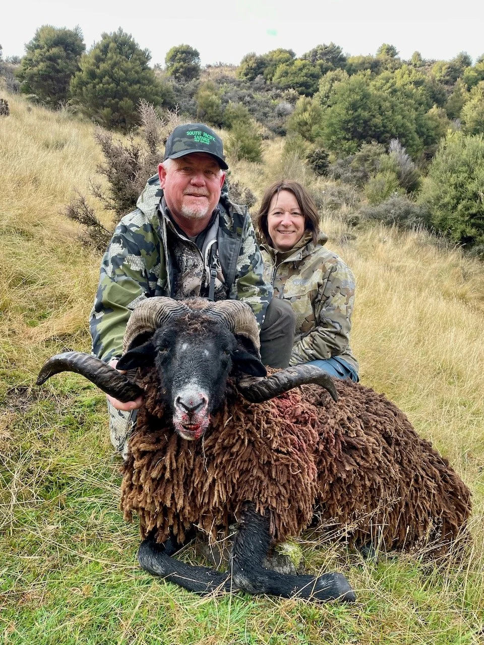 Two hunters, a man and a woman, kneel behind a large, recently hunted ram with curved horns in a grassy field with bushes and trees in the background.