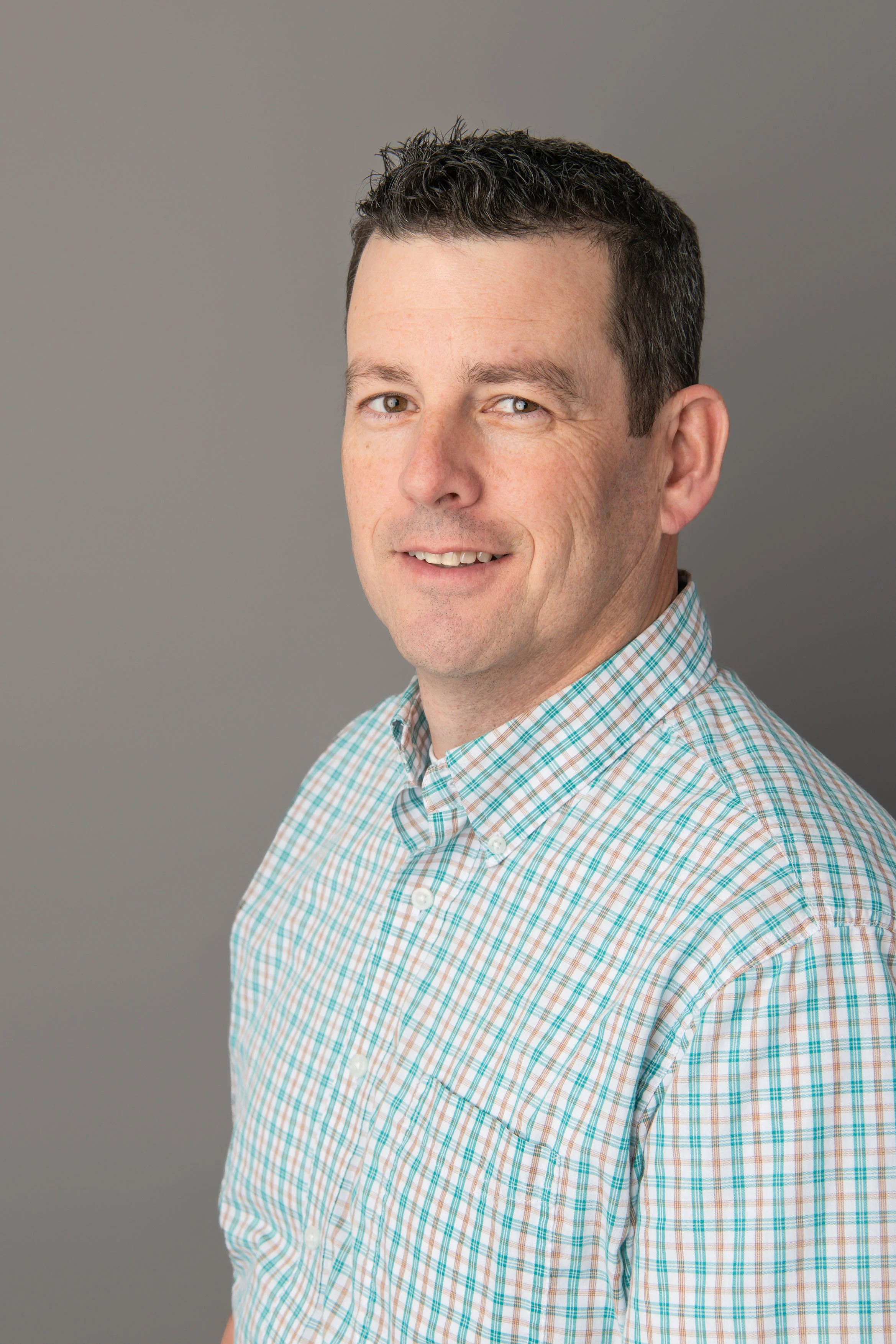 Headshot of a man with short dark hair, wearing a checkered shirt, smiling against a gray background.