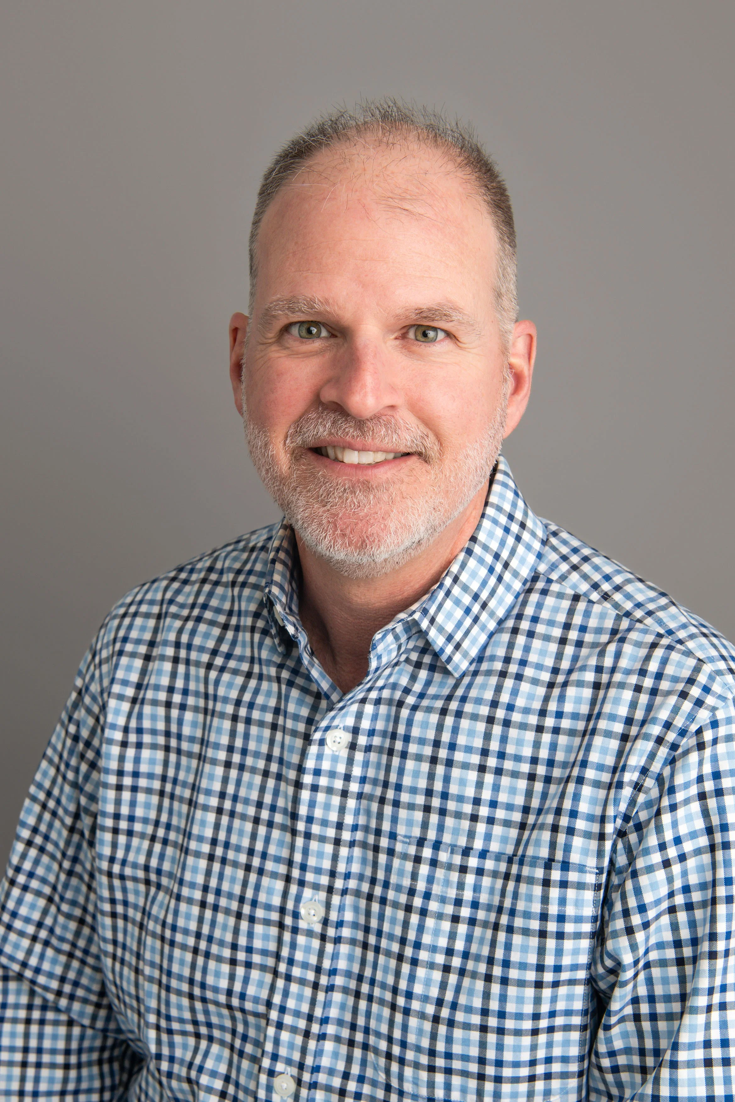 Portrait of a middle-aged man with gray hair and a beard, wearing a blue and white checkered shirt, smiling against a plain gray background.