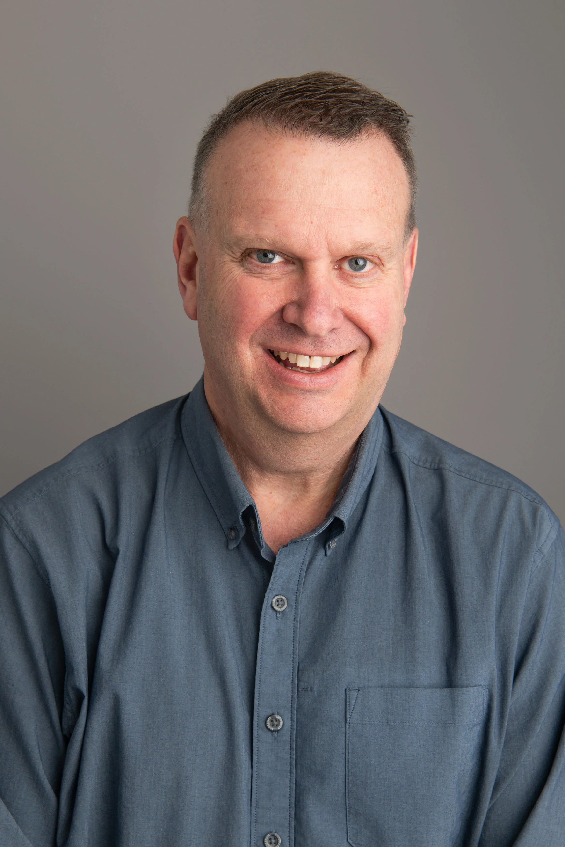 Portrait of a smiling man wearing a blue button-up shirt against a plain gray background.