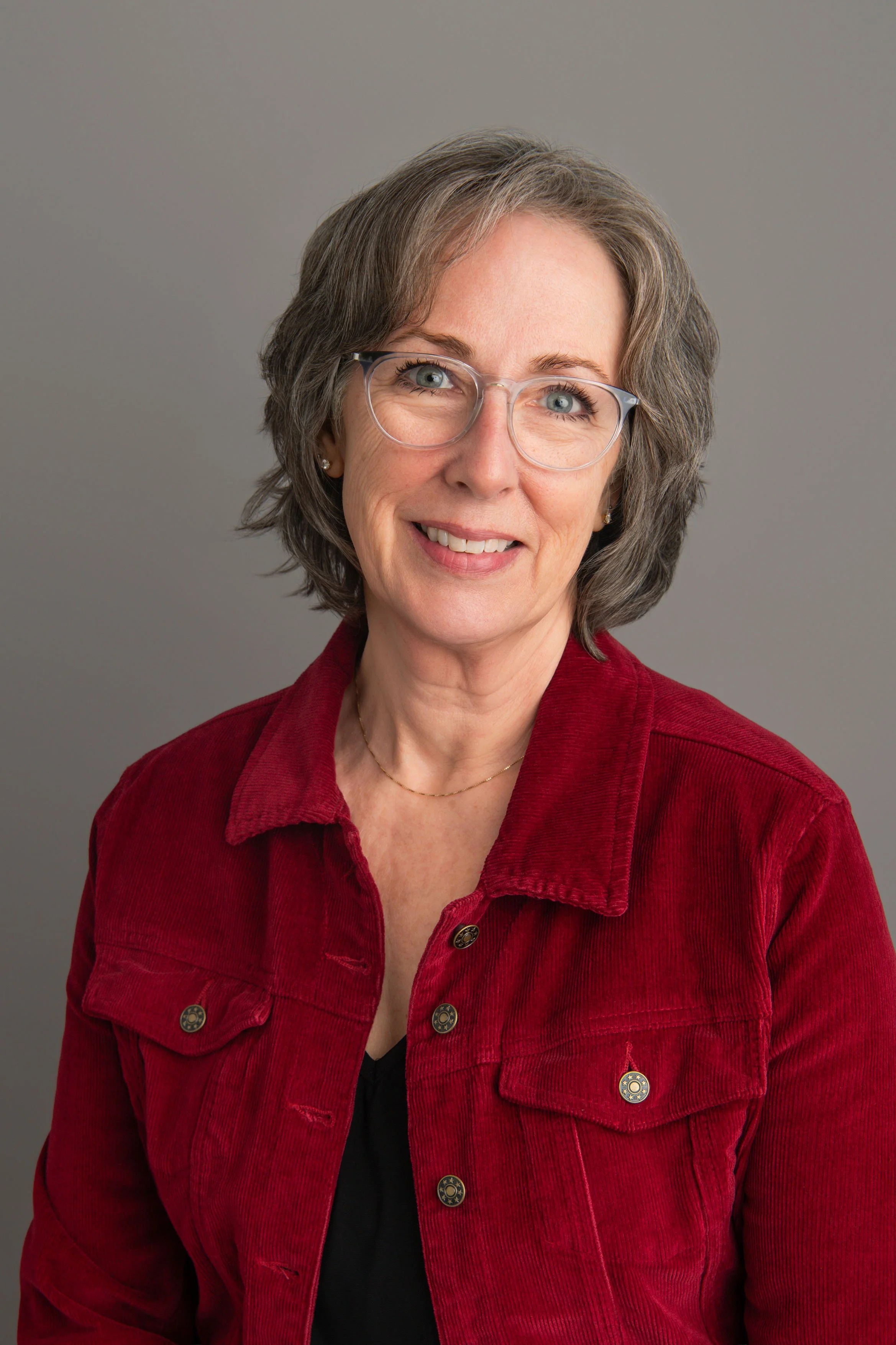 A middle-aged woman with short gray hair, blue eyes, wearing glasses, a black top, and a red corduroy jacket, smiling against a gray background.