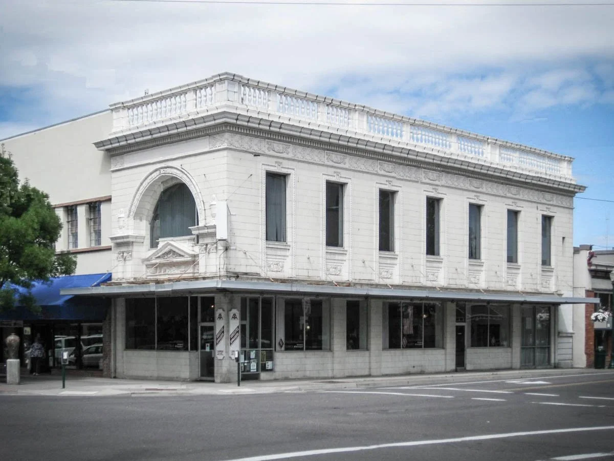 A two-story white building with ornate architectural details, large windows, and a balcony on the upper floor, located on a street corner with a sidewalk and street markings.