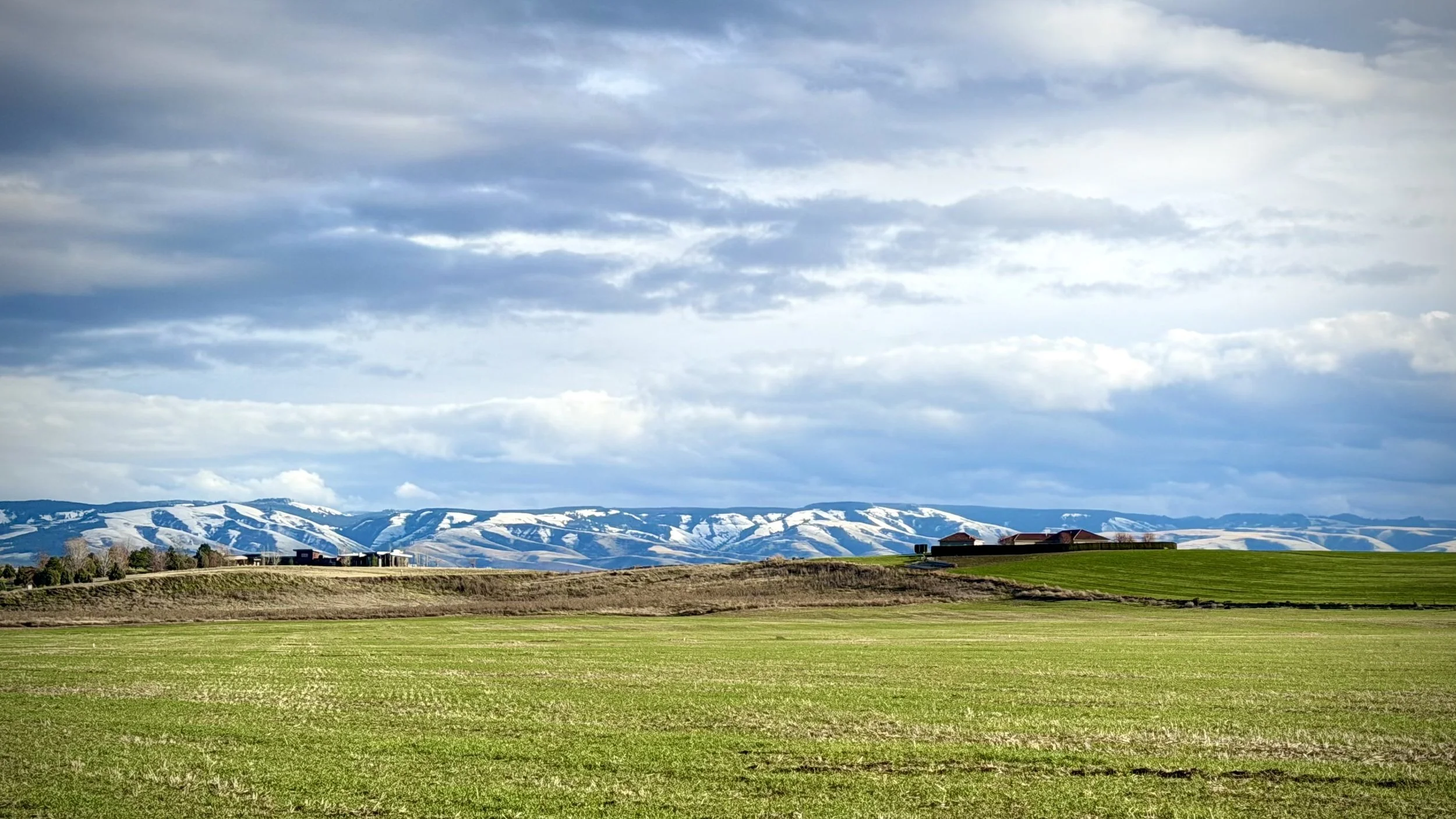 Open field with green grass and distant hills with snow patches under a partly cloudy sky.