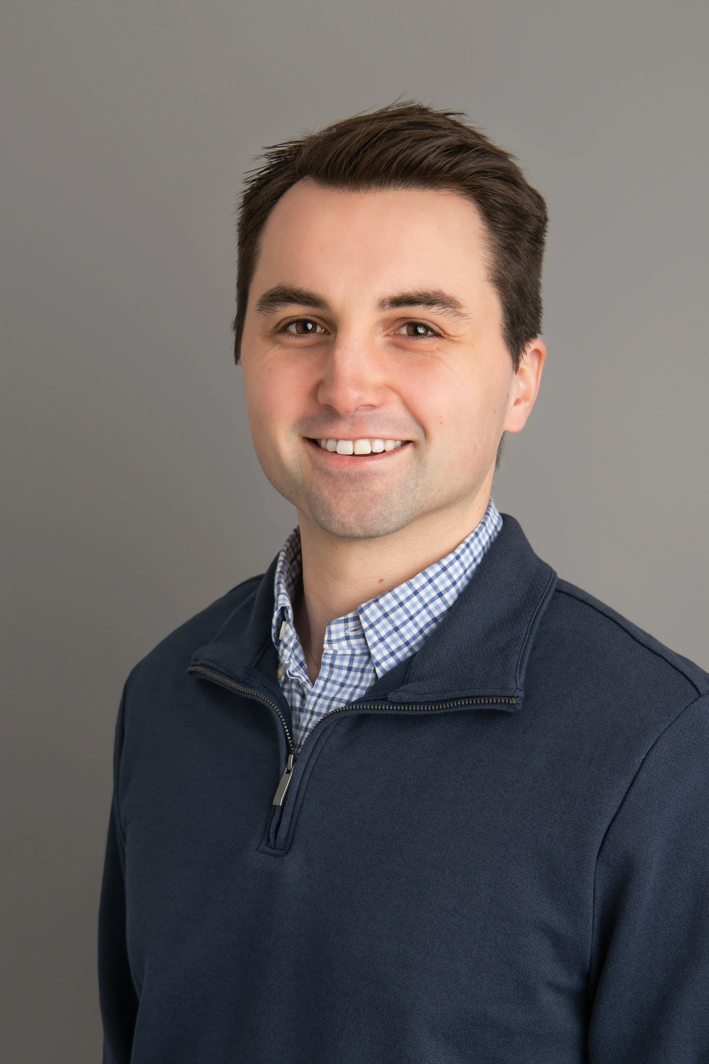 Headshot of a smiling man with short dark hair, wearing a navy blue quarter-zip sweater over a blue checkered collared shirt, against a plain gray background.