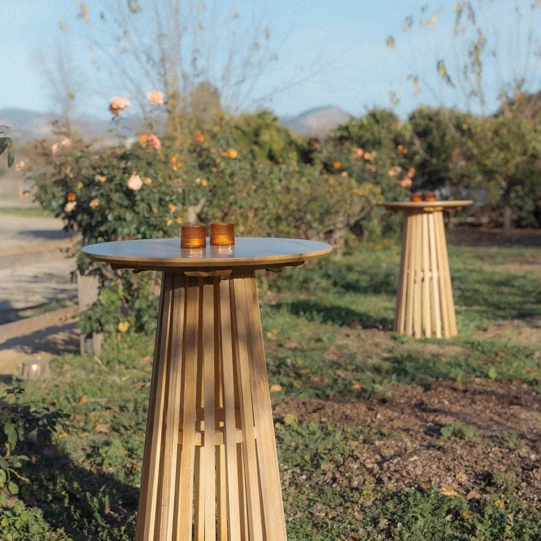 Two wooden outdoor tables with glass cups on top, set in a garden with bushes and trees, in the late afternoon sunlight.