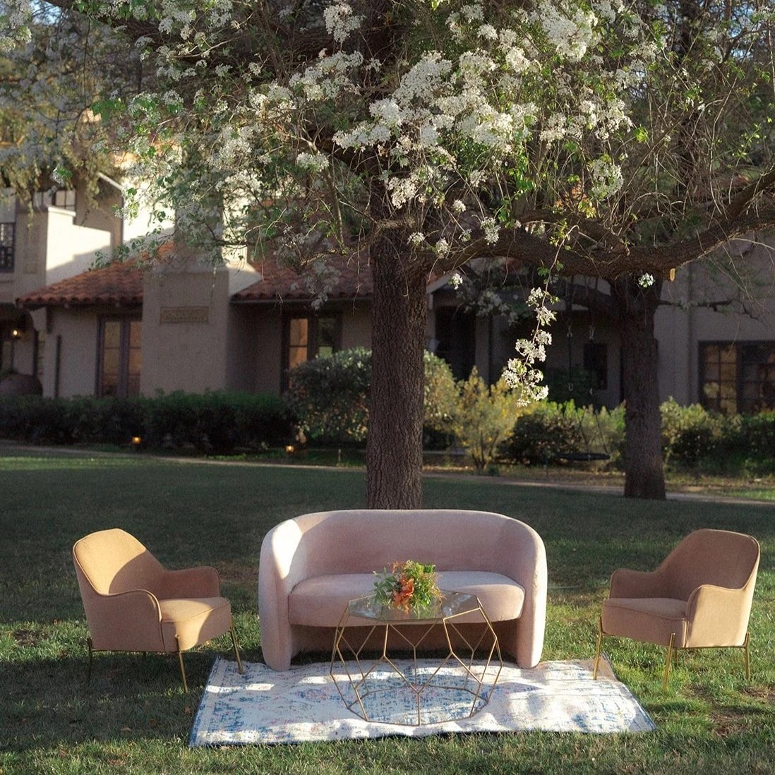 An outdoor seating area with a pink sofa, two matching pink armchairs, a glass coffee table with a floral arrangement, on a patterned rug under a flowering tree with white blossoms, in front of a house with a tiled roof and windows.