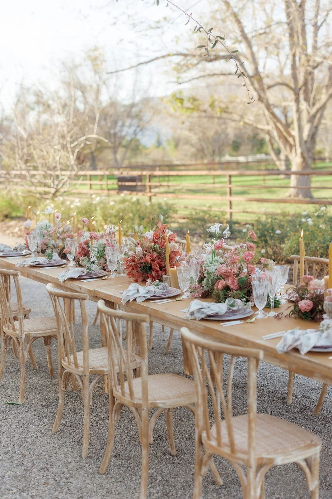 Outdoor wedding or event table decorated with pink and white floral arrangements, candles, and glassware, with a rustic wooden fence and trees in the background.