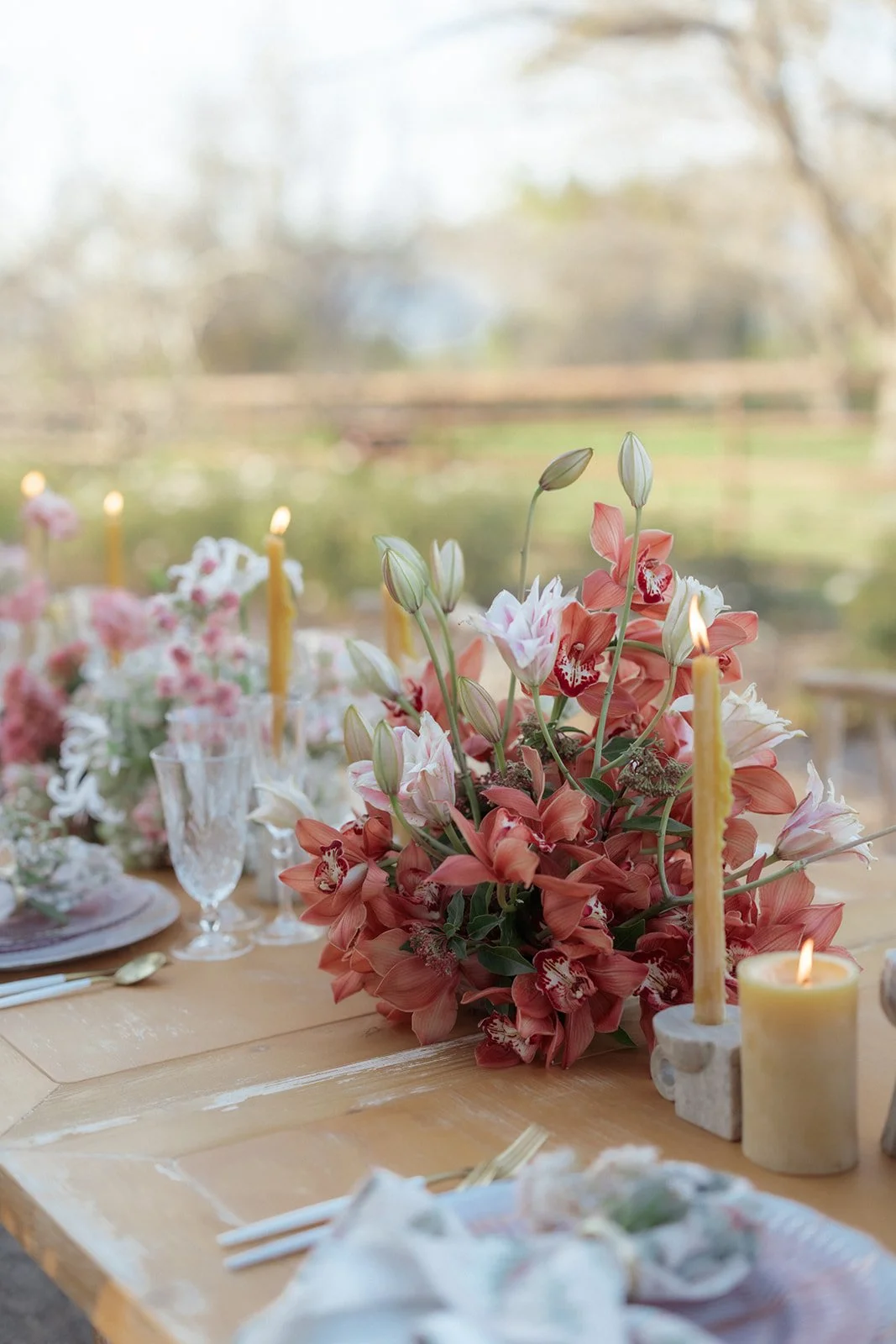 A floral centerpiece with pink orchids on a wooden table, surrounded by candles, glassware, and table settings.