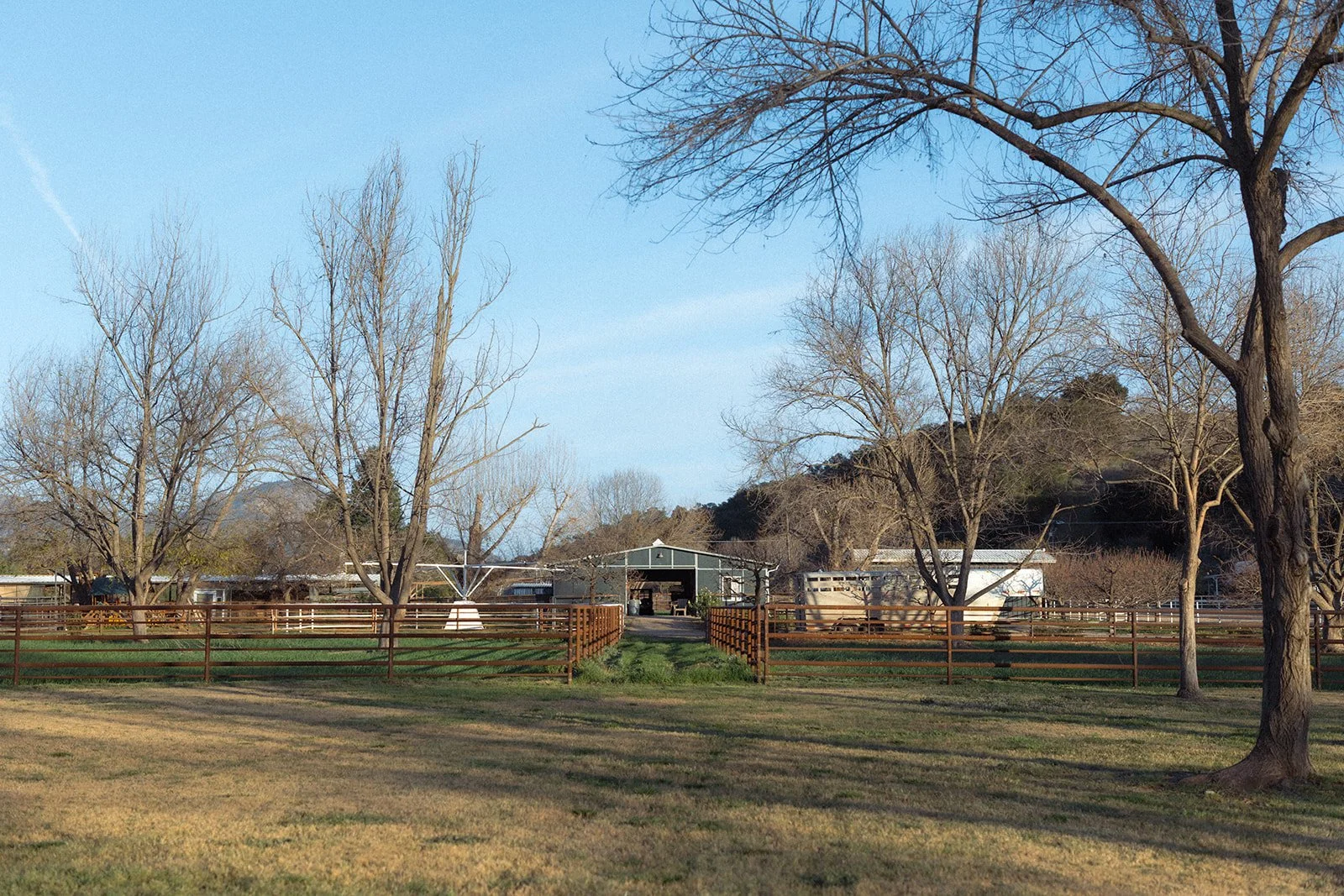 A peaceful farmstead with leafless trees, a wooden fence, a covered barn, and open grassy areas under a clear blue sky.