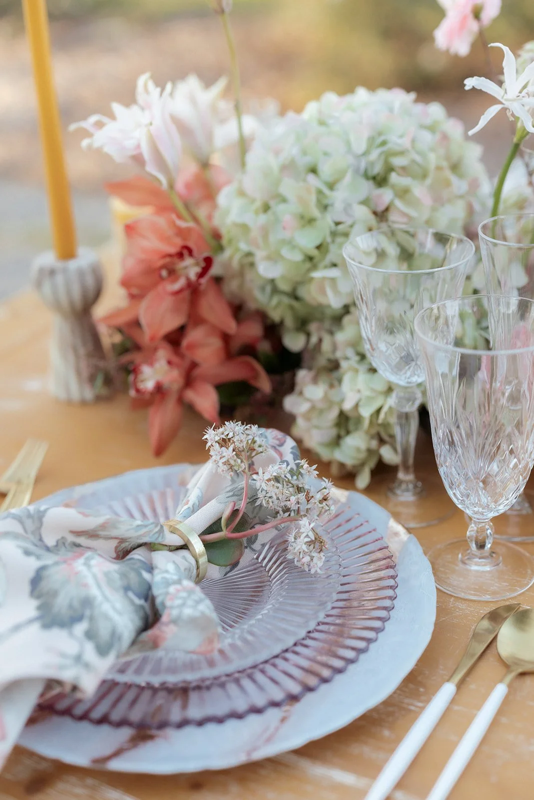 A decorated table with a floral centerpiece, crystal glasses, a plate with a napkin and flowers, gold utensils, and a yellow candle in a holder.