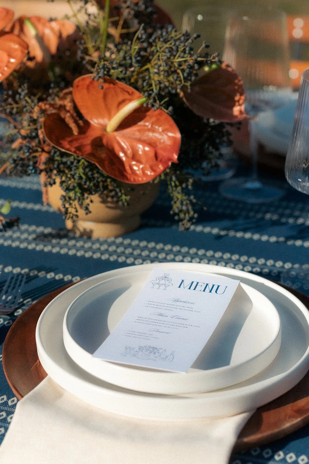 A table setting with white plates, a menu, and a floral centerpiece with brown, orange, and black flowers and greenery, on a blue tablecloth with white dots.